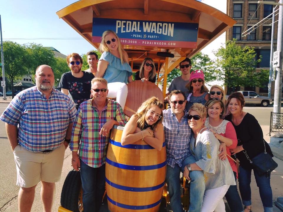 friends family and coworkers smile and pose for a photo during their pedal wagon ride a group of people standing in front of a building